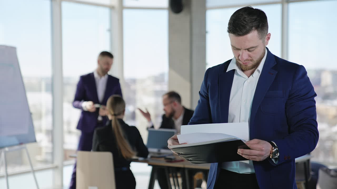 Caucasian man in suit looking through the documents in his hands. Businessman throws the papers emotionally away. Team working at the backdrop in blur.