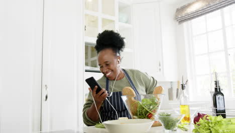 feliz afroamericana anciana bailando con teléfono inteligente y auriculares en la cocina, en cámara lenta