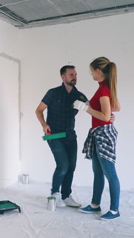 cheerful bearded man paints girlfriend tummy with roller and kisses pretty wife at construction in modern spacious flat