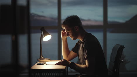 the silhouette of a thoughtful man sitting at a table against the background of a window with oceans and the sea. a restless man sitting at a table with a desk lamp