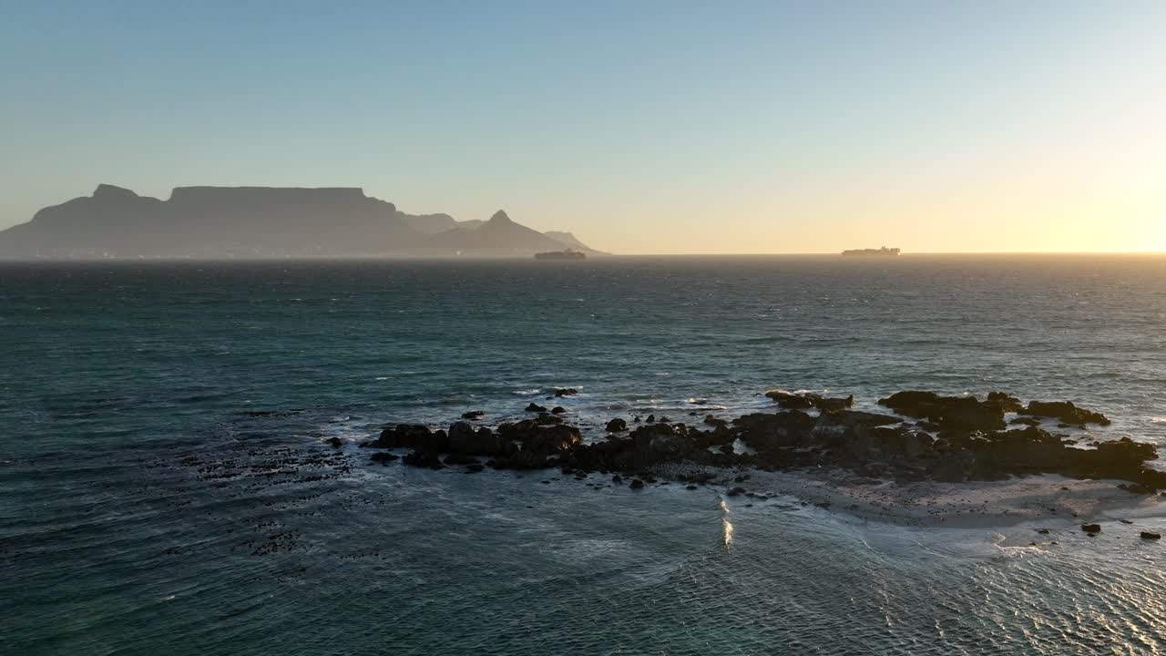 Drone flight above rocky rugged reef with distant sunset view of Table mountain