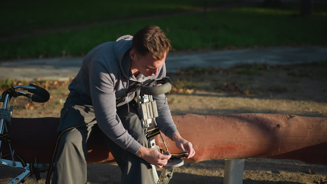 잔디와 개방된 공간으로 평화로운 공원 환경에서 스의 부분을 조정하는 데 집중하여 회색 후디와 바지를 입고 야외에서 로그와 같은 구조에 앉아있는 남자