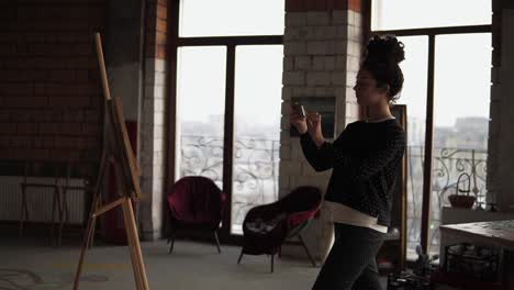 Young-curly-girl-proud-of-herself-and-her-work,-taking-picture-on-her-smartphone-of-the-canvas-with-painting-she-drew.