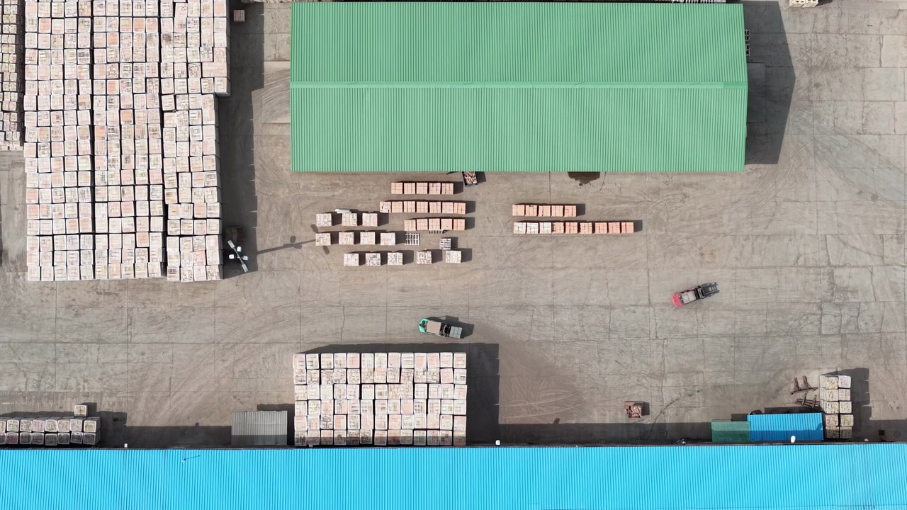 An aerial drone shot of a busy industrial complex displays massive goods stacks and active forklifts on a concrete yard, depicting vital logistics operations.