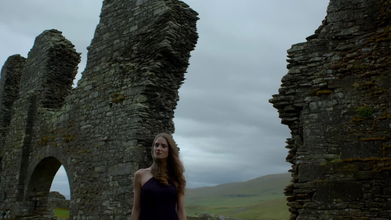 Woman stands among ancient stone ruins under a cloudy sky