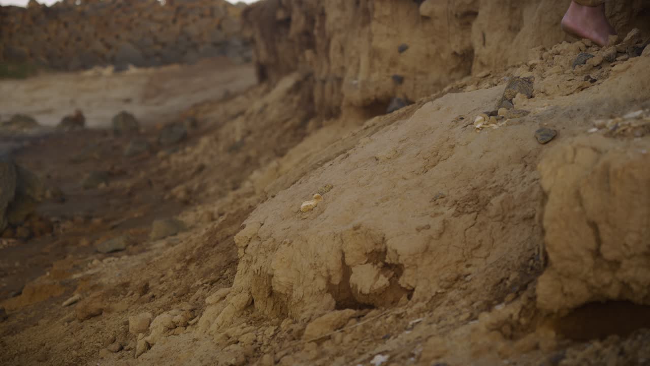 A barefoot individual ascends a rugged sand-covered incline by the coast, hands steadying against the earth, symbolizing resilience, grounding, and human connection to nature’s raw landscape