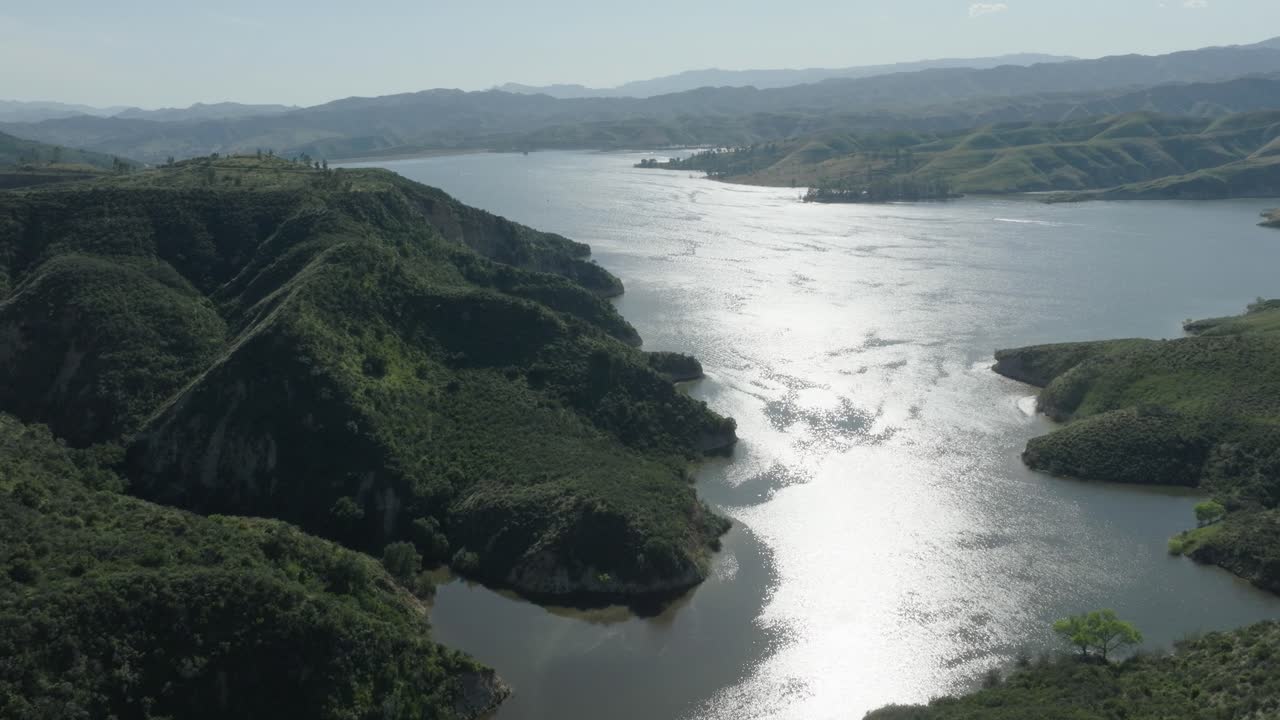 Aerial pull-back shot showing the shimmering surface of Castaic Lake in Santa Clarita, Ca. on a hazy sunny day with lush green hillsides and small watercraft on the water.