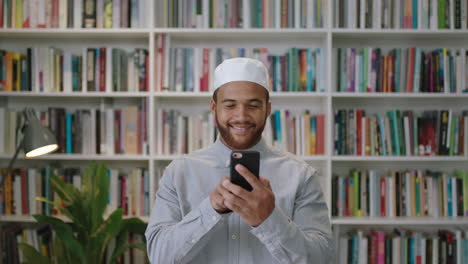 Joven-Hombre-Confiado-Del-Medio-Oriente-Parado-En-La-Biblioteca-Usando-Un-Teléfono-Inteligente-Retrato-Sonriente-De-Un-Orgulloso-Empresario-Usando-Las-Redes-Sociales-Riéndose