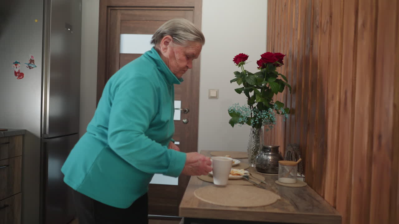 Senior woman in green fleece holding cup, preparing for breakfast. Cozy kitchen setting with roses on table, plates with bread, tea cup, and natural light creating peaceful atmosphere