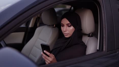 attractive young woman in black hijab sitting in her car, thoughtfully looking at her smartphone screen