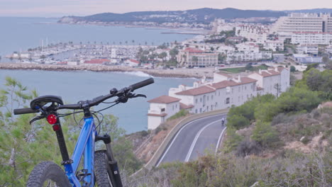 Ein-Geparktes-Mountainbike-Blickt-Auf-Die-Atemberaubende-Aussicht-Auf-Die-Küste,-Während-Radfahrer-Und-Autos-Auf-Der-Kurvenreichen-Straße-Darunter-Navigieren