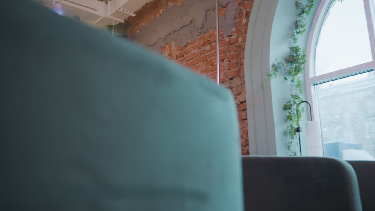 Lady with long hair walks forward in modern indoor space, reaching out to touch leaf of potted plant beside arched window, surrounded by green plants, natural daylight, and cozy furnishings