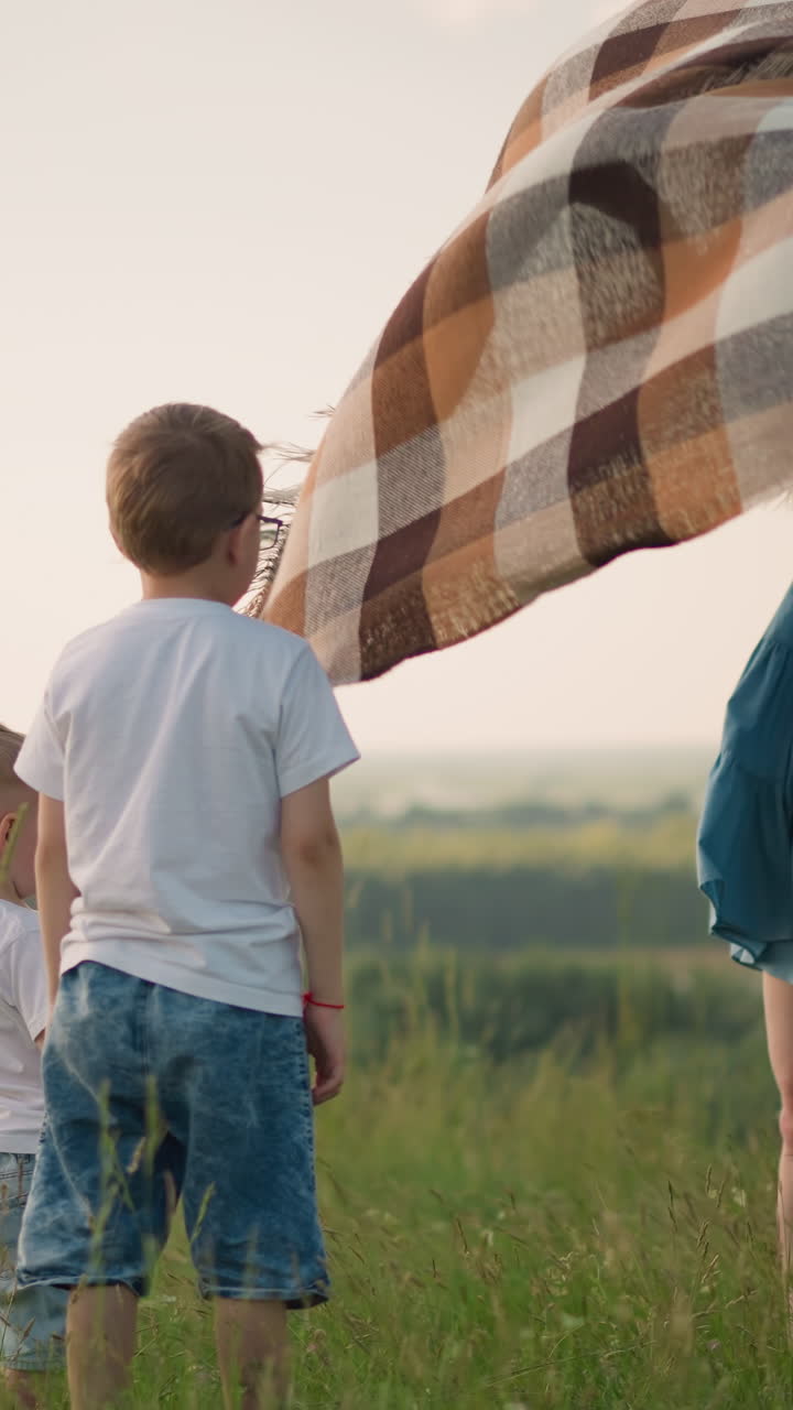 A woman in a blue dress holds a large plaid scarf high as it catches the wind on a lush grassy hilltop. Two children in white shirts stand in front of her, observing the flowing scarf, all under a sky