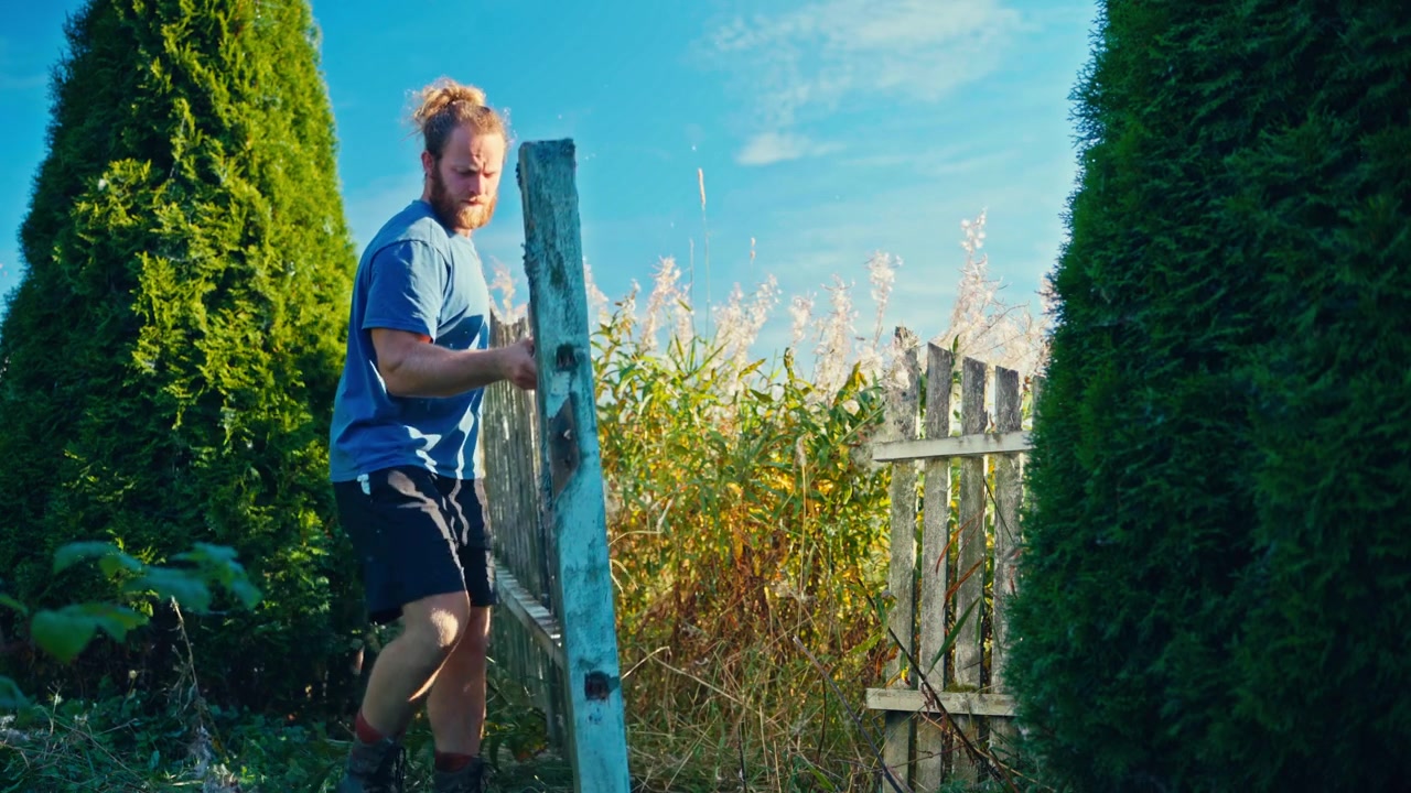 Un homme démonte une clôture en bois dans un jardin, entouré de verdure et de lumière du soleil - Plan fixe