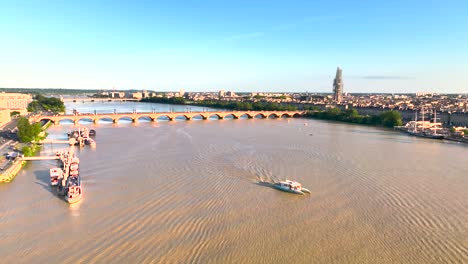 Garonne-River-with-boats-and-large-sailboats-right-with-Saint-Michel-Basilica-Bell-tower-in-scaffolding,-Aerial-flyover-shot