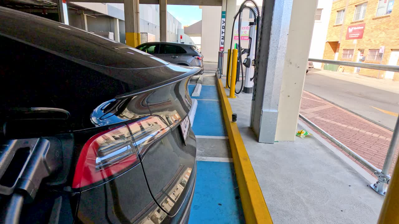 Un'auto elettrica nera si ricarica presso una stazione pubblica mostrando lo stato della batteria sullo schermo.