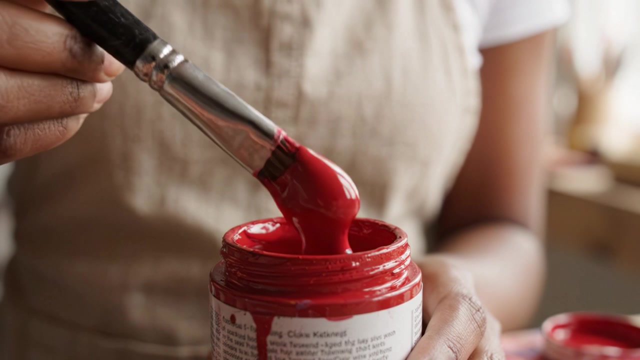 Un primo piano di un artista che tiene un pennello intinto in vernice rossa vibrante sopra un barattolo di pittura, mentre si prepara per un progetto creativo al chiuso.