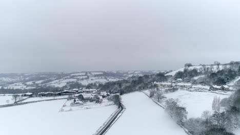 Aerial-tracking-sideways-across-snow-covered-fields-and-farmland-buildings-and-dwellings,-Devon,-UK