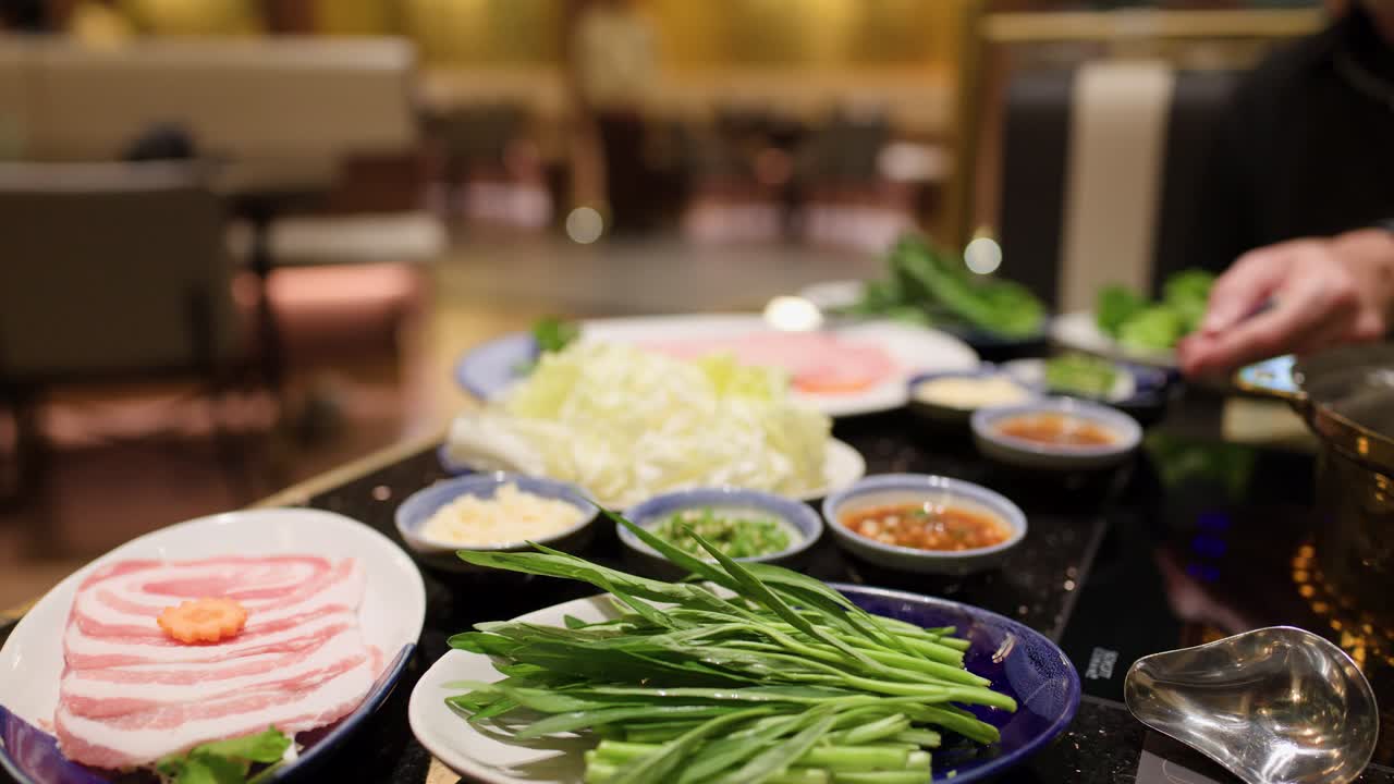 Hand places vegetables into hotpot with sliced meat, sauces, and greens under warm indoor lighting