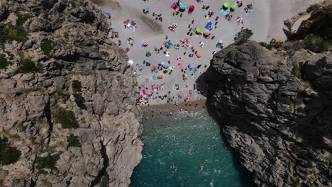 Aerial-drone-Shot-above-the-remote-beach-at-Torrent-de-Pareis-where-towering-cliffs-surround-a-stony-beach-and-multiple-boats-anchored-up