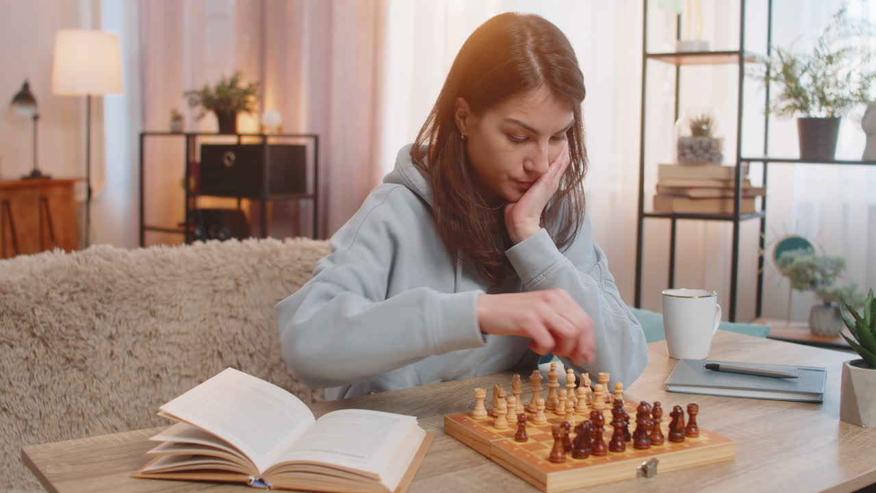 joven jugando al ajedrez sola arreglando piezas en la mesa en el sofá con el libro acostado cerca en casa