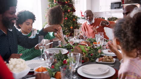 mixed race, multi generation family sitting at their christmas dinner table serving themselves food and talking together, selective focus