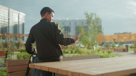 young teen outdoors retrieves book from backpack and drops it onto wooden table, urban background with greenery and buildings visible under natural daylight