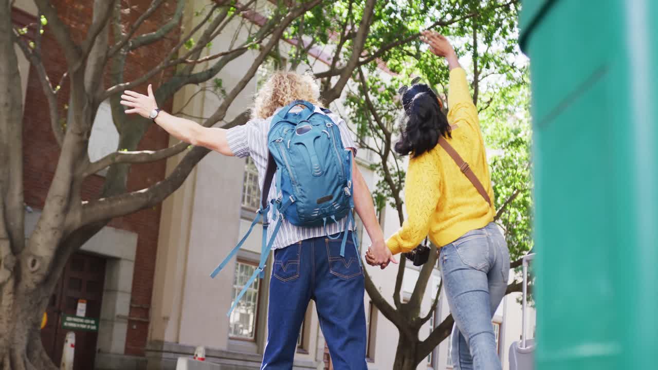 vista posterior de una pareja diversa con equipaje llamando a un taxi en la calle de la ciudad