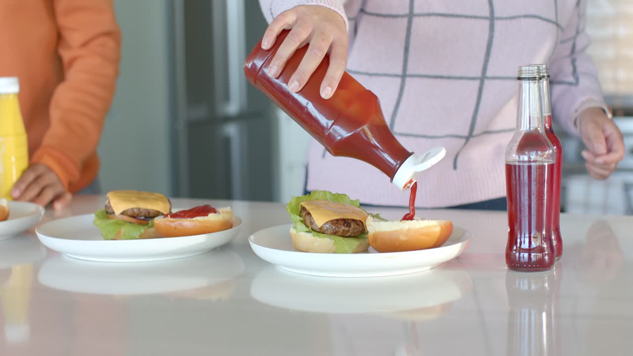 Amici al bancone della cucina, con un berretto in mano, una donna che mette il ketchup sul panino e un uomo che prende la senape