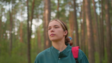 lady wearing green shirt with red backpack walks along a forest trail surrounded by tall trees, gazing thoughtfully with her friend blurred in background, wearing a blue scarf and looking unhappy