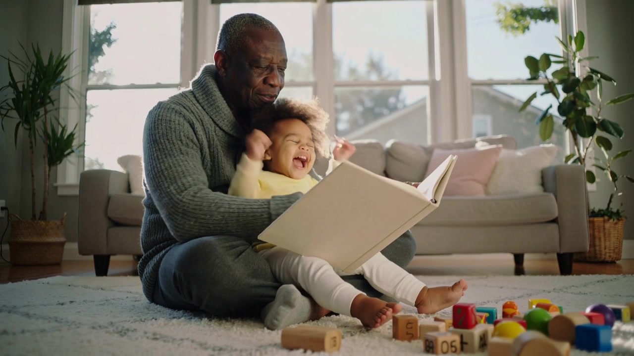 Nonno in maglione grigio che apre un libro illustrato e gira le pagine con un bambino piccolo sul tappeto, condividendo una storia.