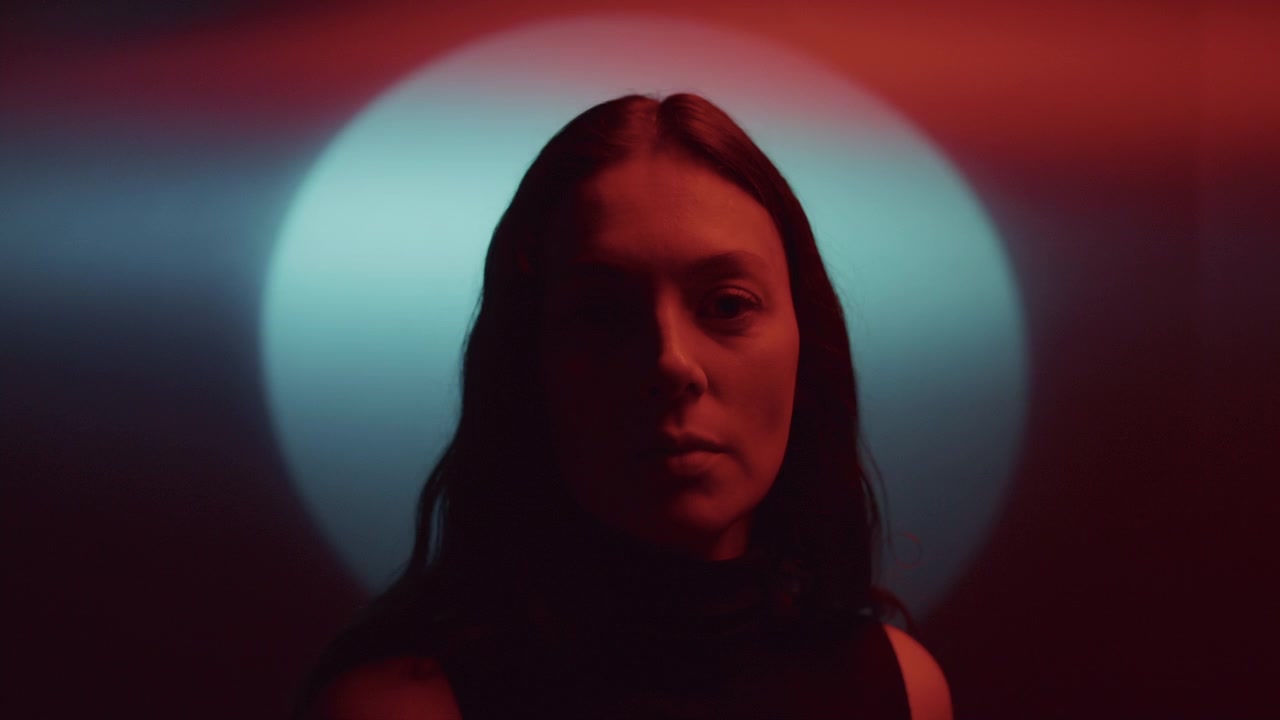 Ritratto in studio di donna bruna illuminata da luce rossa contro un cerchio blu luminoso.