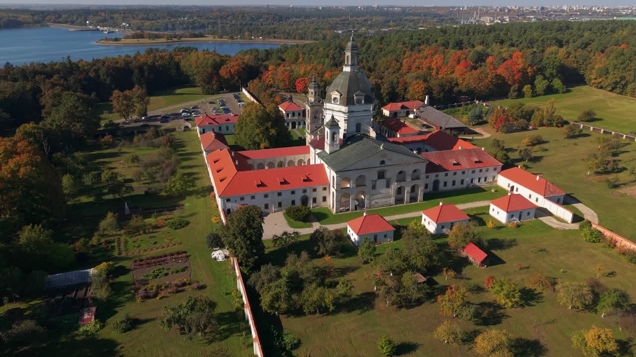 Aerial view of Pažaislis Monastery in Kaunas, Lithuania, surrounded by vibrant autumn foliage and forest, with the Baroque architecture standing out under clear blue skies