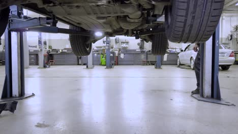 Mecánico-De-Automóviles-Trabajando-En-El-Automóvil-Bajándolo-En-El-Ascensor-En-El-Taller-De-Automóviles-De-Alto-Nivel