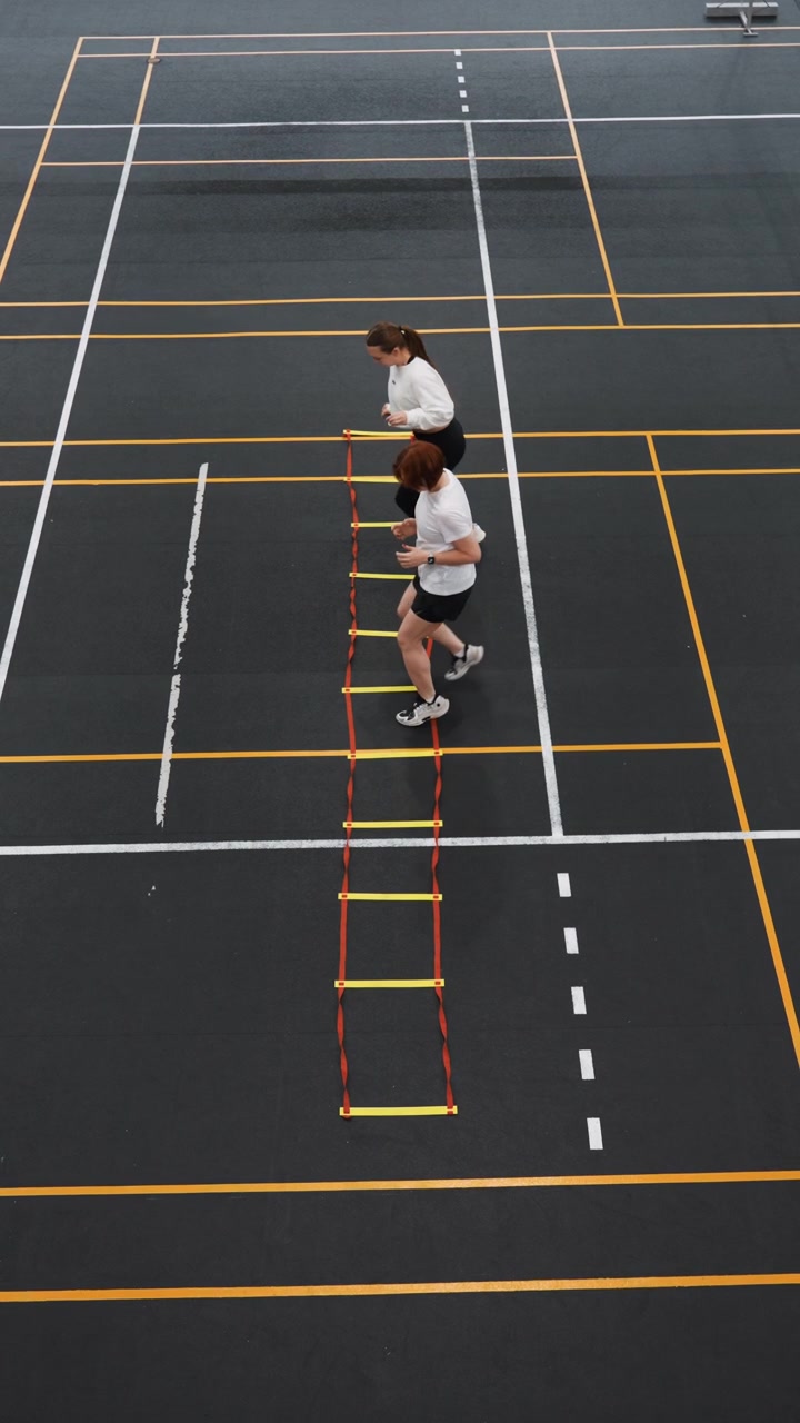 Overhead gym court paired ladder drills. Two athletes run sidebyside through rungs under instructor supervision, rapid exchanges, synchronized steps, collaborative competition vibe, intense