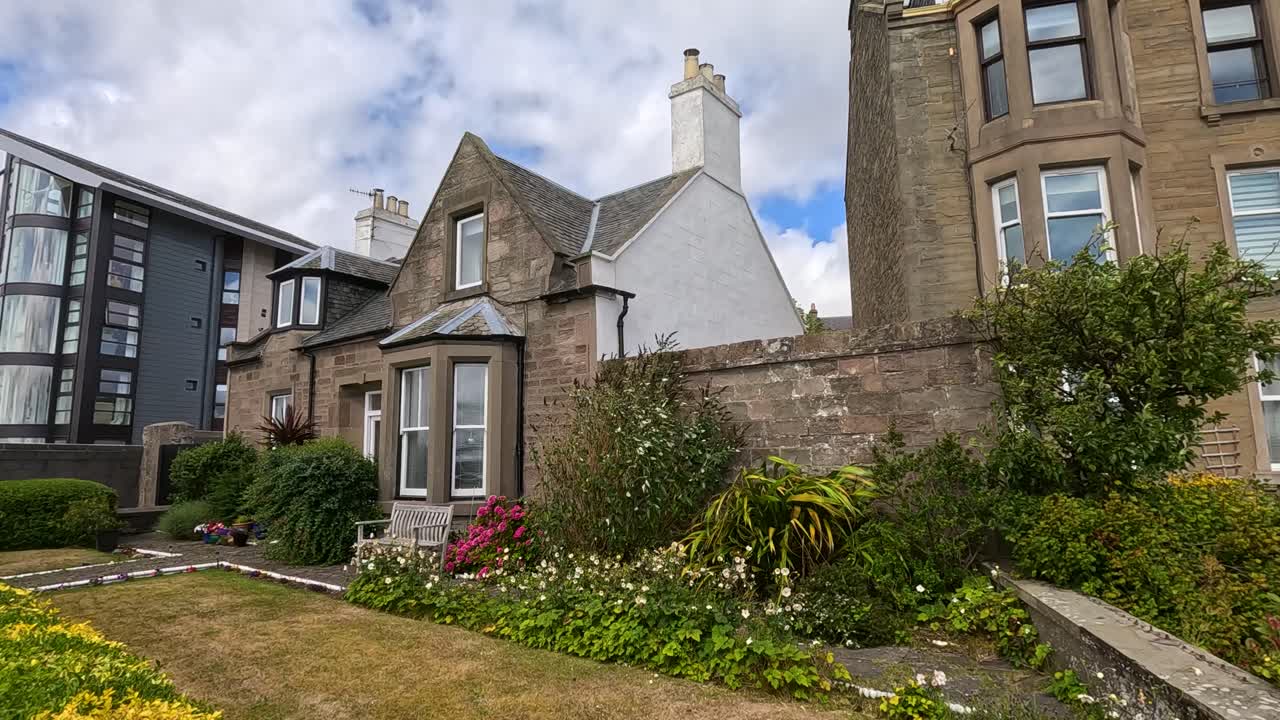 Camera slowly pans across a traditional stone house with lush garden in Broughty Ferry, Dundee, under bright daylight and partly cloudy skies