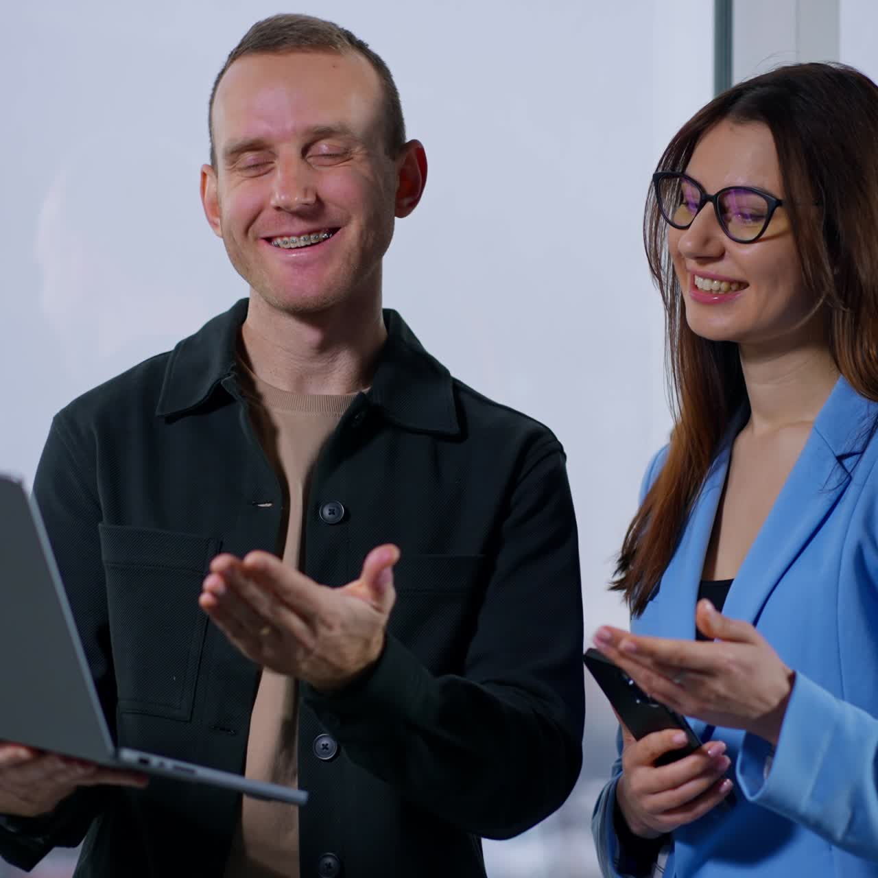 Man and woman working in the office stand at the panoramic window. Smiling colleagues look at laptop and point at it with hand
