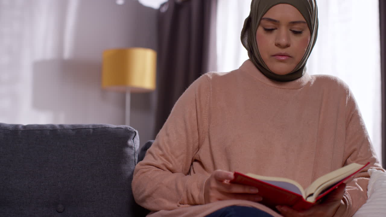 Donna musulmana con l'hijab seduta sul divano a casa che legge o studia il Corano
