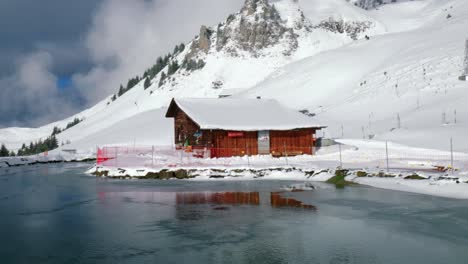 drone panning from the right to the left side of the frame, showing a frozen lake in front of a log cabin at a snowy mountainside of the alps in engelberg, switzerland