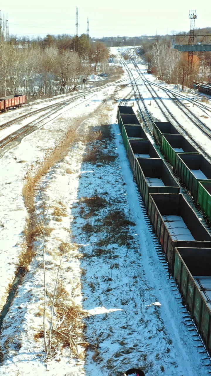 A freight train pulls a trailer with containers behind on the railroad in the background of the square of rail station with a multitude rows of container on the outskirts of the city in the winter. Aerial view. Vertical video