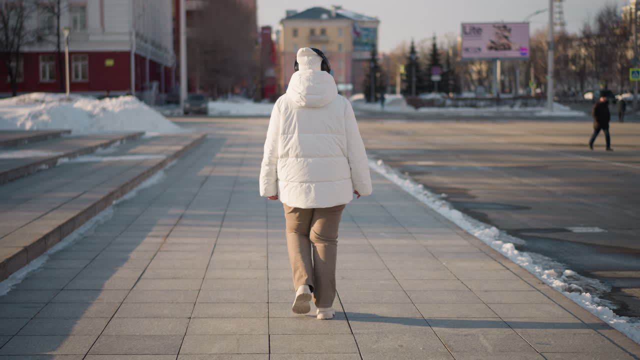 Back view of woman walking down tiled path beside steps on busy urban street during winter, wearing puffy coat and beanie, snow piles and billboard, cars and pedestrians in background