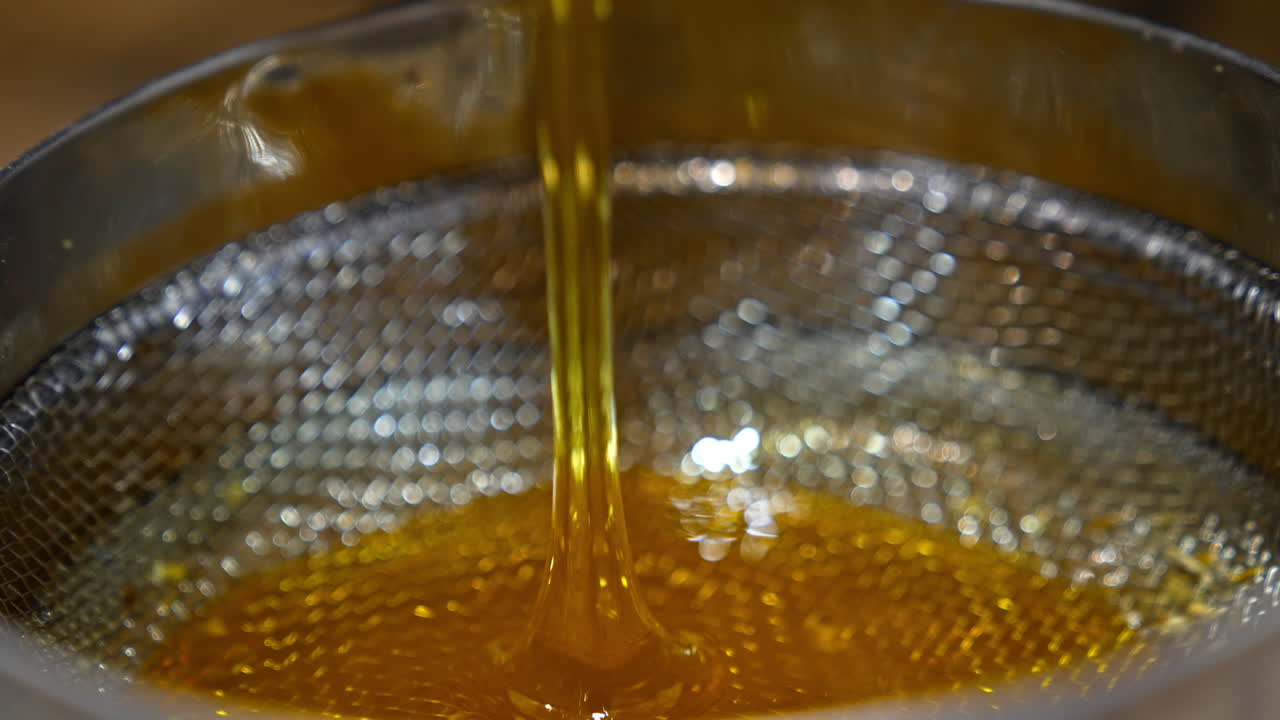 Little stream of honey flowing onto the metal sieve. Close up. Organic honey filtering after being extracted from frames.