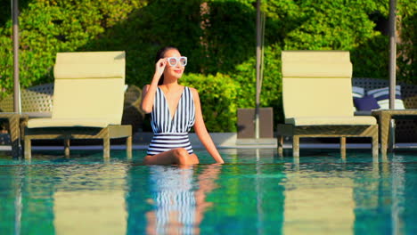encantadora mujer asiática con traje de baño blanco y negro sentada en el agua de la piscina