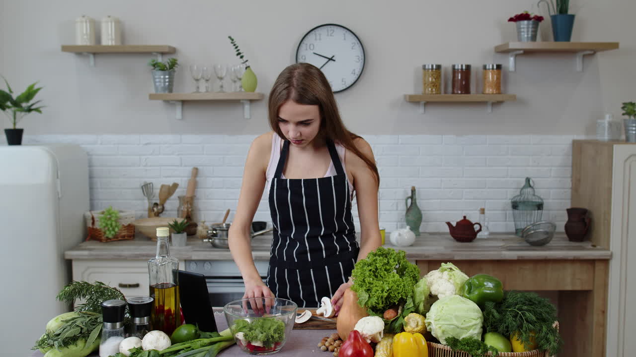 Niña vegana cocinando ensalada con verduras crudas mientras mira en una tableta digital la receta en línea