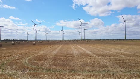 Das-Luftvideo-Führt-Uns-über-Lincolnshire,-Wo-Sich-Windkraftanlagen-Anmutig-Auf-Dem-Gerade-Abgeernteten-Feld-Eines-Bauern-Drehen,-Umrahmt-Von-Der-Warmen-Präsenz-Goldener-Heuballen