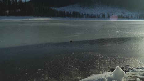 Frozen-mountain-lake-in-winter-with-snow-on-shoreline-and-evergreen-trees-on-opposite-shore