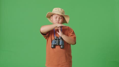 asian tourist boy with a hat and binoculars shaking head showing time out gesture on the green screen background. boy researcher, travel tourism adventure concept