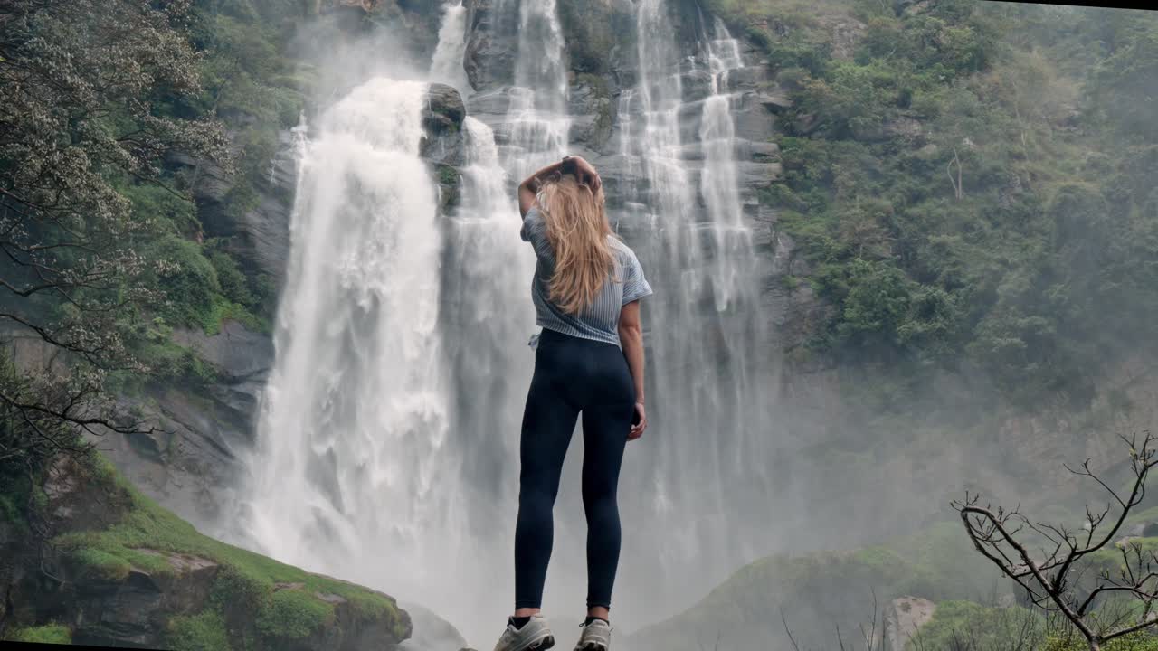 A woman stands in awe before the roaring Bomburu Ella Waterfalls, surrounded by mist, jungle, and rocky cliffs in the lush highlands of Nuwara Eliya, Sri Lanka.