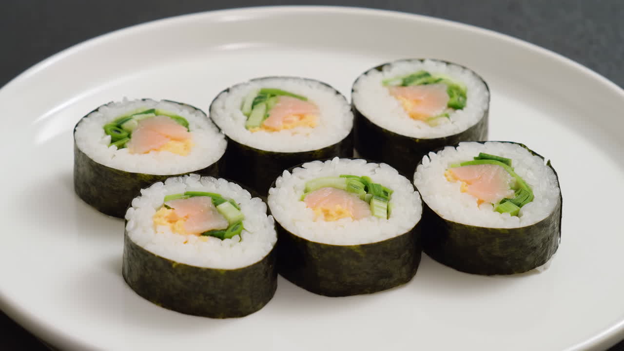 Gulungan Sushi di Piring Putih Menampilkan Salmon, Mentimun, dan Nori dalam Susunan Rapi dengan Pencahayaan Studio yang Lembut.