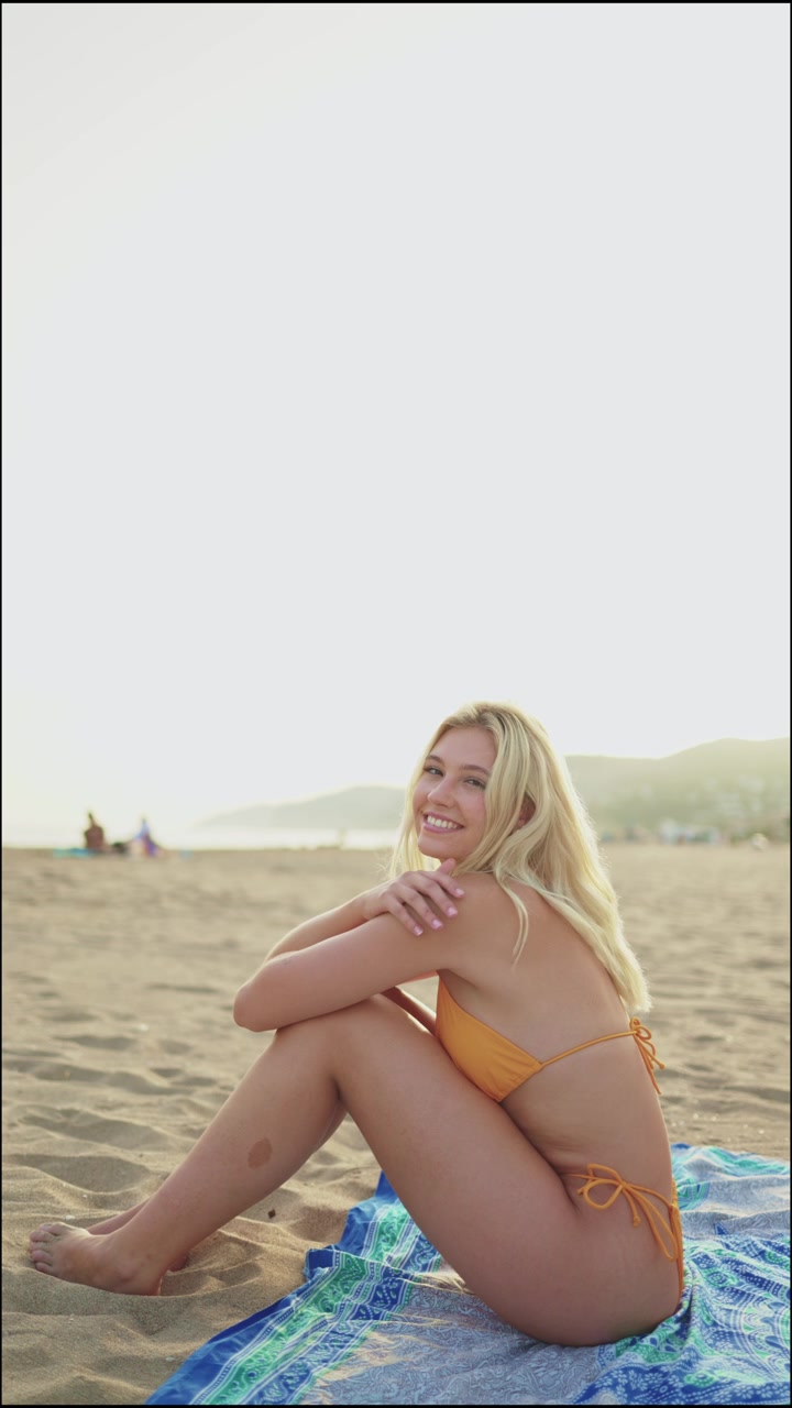 Donna bionda felice in bikini seduta su una spiaggia sabbiosa.